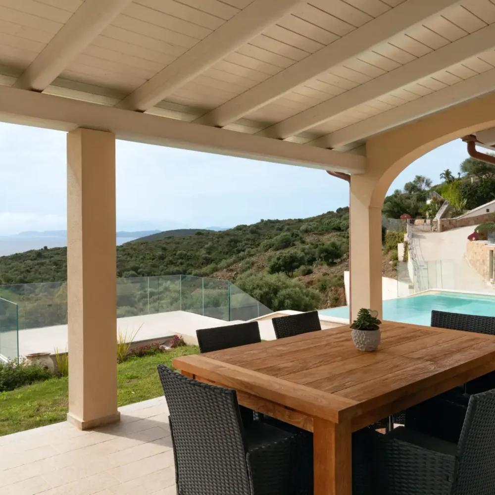 Covered terrace with wooden dining table and sea view, overlooking a private pool and green hills